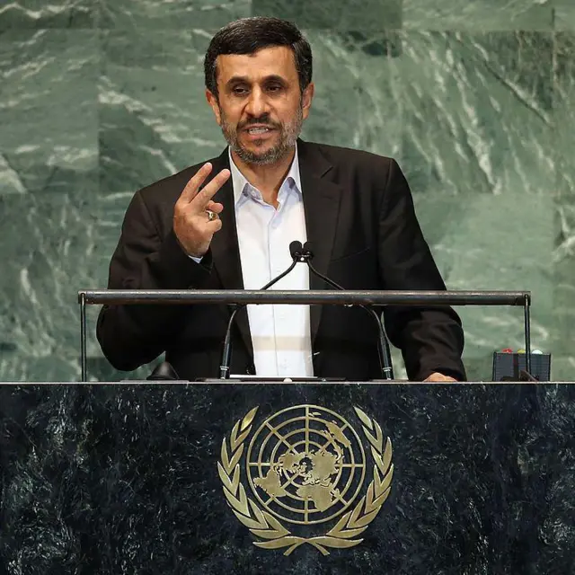 Mahmoud Ahmadinejad habla de pie en el podio de oradores de la ONU. Viste una chaqueta oscura y una camisa clara sin corbata. Apoya su mano izquierda sobre la mesa y tiene levantada la mano derecha a la altura del pecho mostrando dos dedos.