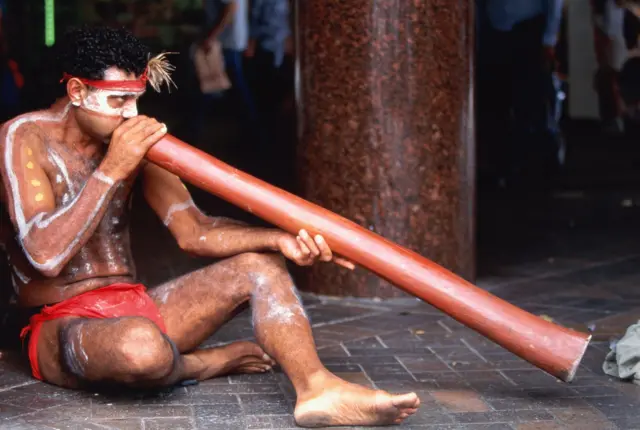 Un aborigen australiano, en taparrabos rojo,  con la cara y cuerpo pintados de líneas y puntos blancos, está sentado soplando por un tubo largo de caña