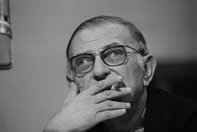 Sartre, con sus lentes, fumando un cigarrillos que tiene entre los dedos