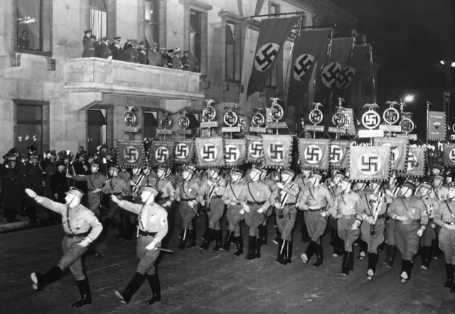 Soldados con uniformes y botas negras desfilan y llevan estandartes que tienen la esvástica