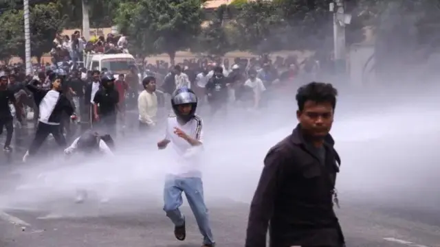 Un hombre con casco de motocicleta, pantalones azules y camiseta blanca corre bajo un chorro de gas o agua mientras la multitud lo sigue, el 8 de septiembre de 2025. Un hombre levanta el brazo, como si lanzara un objeto. Un hombre en primer plano mira a la cámara.