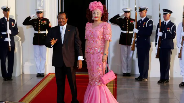 El presidente de Camerún con traje gris y corbata blanca, saluda a los fotógrafos. Su esposa, Chantal, con un vestido rosa floreado y un tocado haciendo juego, está junto a él.