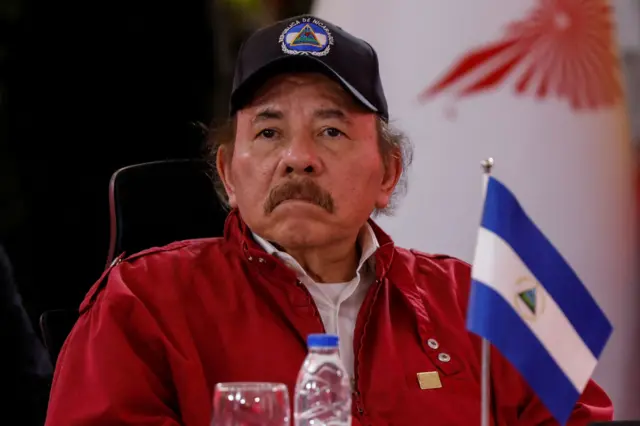 El líder de Nicaragua Daniel Ortega, sentado con una cachucha con el escudo de NIcaragua, una chamarra roja y frente a él, una banderita de Nicaragua