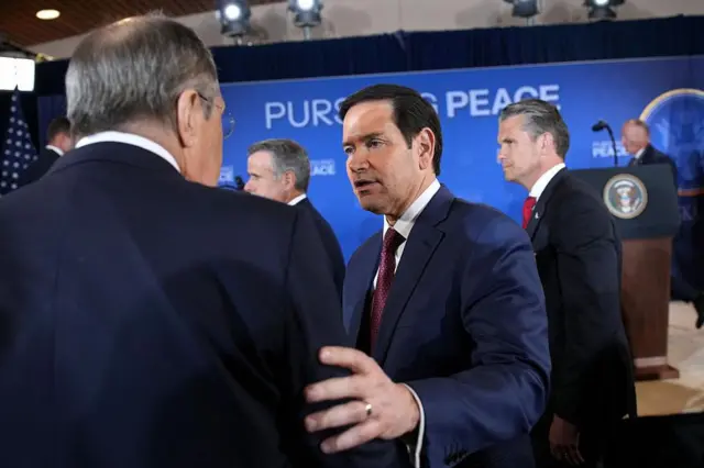 Lavrov está de espaldas a la cámara; Rubio le dice algo y le toca el hombro con la mano. El secretario de Defensa de Estados Unidos, Pete Hegseth, está detrás de ellos, frente a un cartel que dice 