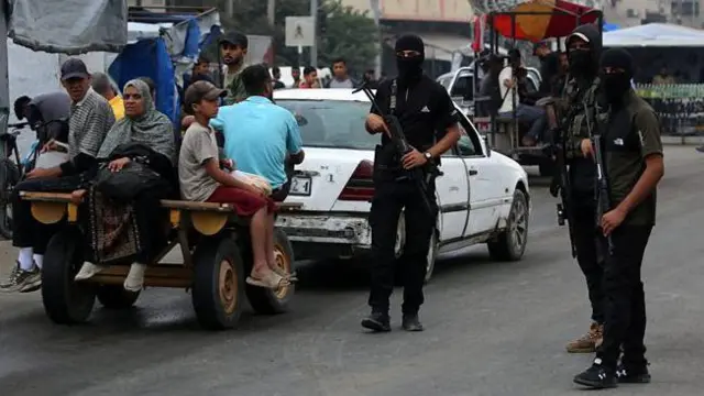 Tres milicianos con el rostro cubierto y armados con fusiles patrullan una calle en Gaza. Junto a ellos, pasa un auto blacno que tira de un carro con una mujer y un niño.