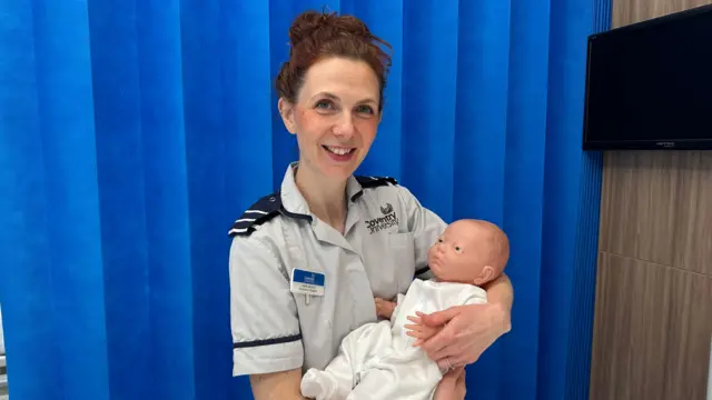 Una mujer con pelo castaño recogido, vestida en uniforme de enfermera, sotiene a un bebé muñeco