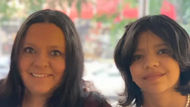 Cynthia Peralta, una mujer de cabello castaño largo, sonríe a la cámara mientras posa para una foto con su hija, Juliana, quien lleva un collar de peras y también sonríe.