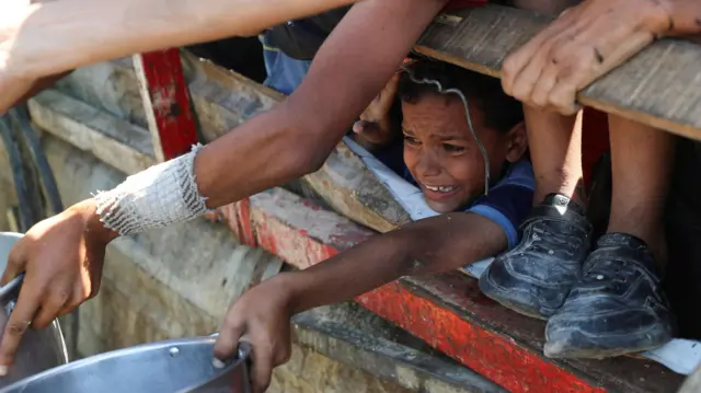 Un niño espera en una valla con su cazo preparado para recibir algún tipo de comida.