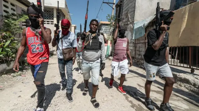 Cinco jóvenes con pantalones cortos y jeans rotos caminan por una calle sin pavimentar con armas en alto mientras patrullan una zona de Puerto Príncipe en febrero de 2024.