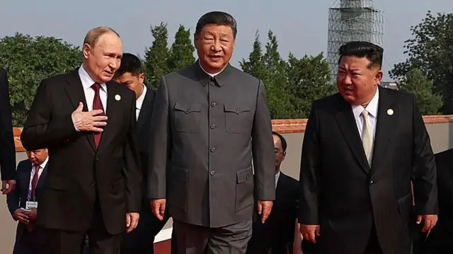 Vladimir Putin, Xi Jinping y Kim Jong-un, de pie, hombro con hombro, todos con traje, con otros con traje detrás. Putin se lleva la mano derecha al pecho.