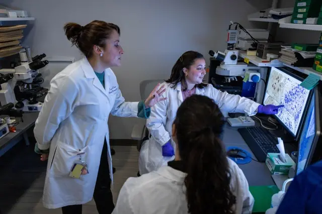 Tamar Gefen, de pie, ve unas imágenes en una pantalla de una computadora. Dos investigadoras están sentadas y una de ellas está señalando un punto en la pantalla. Las tres llevan batas de laboratorio y están en una oficina con equipos médicos
