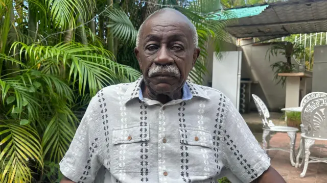 Un hombre anciano, de bigote, viste una guayabera gris de manga corta y mira directo a cámara 