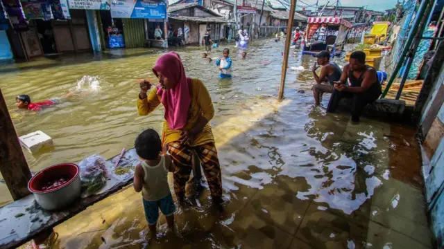 Adultos sentados con los pies sumergidos en el agua al borde de la carretera, mientras que los niños vadean y nadan por la calle inundada en el norte de Yakarta, noviembre de 2024.