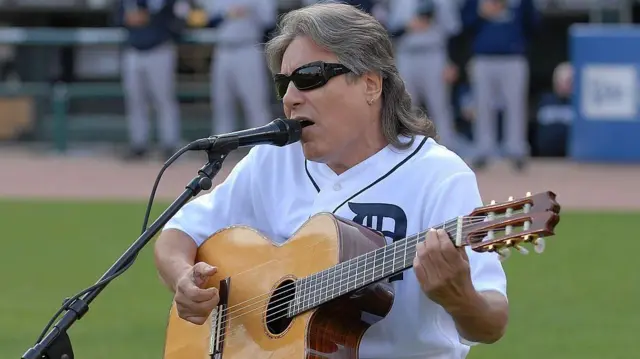 Feliciano interpreta el himno de EE.UU. antes del juego de los Tigres de Detroit y los Yankees de Nueva York en el Comernica Park en Detroit, Michigan. Está sentado frente a un micrófono en medio del estadio, sostiene su guitarra, lleva sus gafas negras en la cara y el cabello hasta los hombros. La imagen es de 2010. 