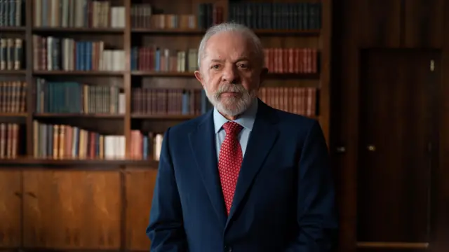 Lula de pie. Vestido con traje azul oscuro, camisa clara y corbata roja mira hacia la cámara. En el fondo borroso, se aprecia una biblioteca llena de libros.