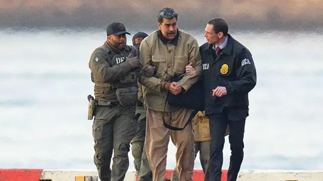 Nicolás Maduro in handcuffs wearing a beige coat and trousers after landing at a Manhattan helipad. Two men hold his arms and two more people follow behind.