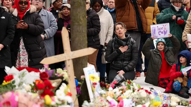 Residentes de Mineápolis en la calle donde Renee Good fue baleada. Se ve a adultos y niños. Una mujer arrodillada expresa su dolor, mientras se ve en el suelo numerosos ramos de flores y una cruz de madera