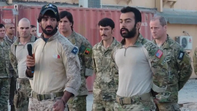 Safi de pie en posición militar junto a sus compañeros. Lleva un uniforme militar y está de pie con las manos a la espalda. Cinco compañeros están de pie detrás de él y otro está a su lado sosteniendo un micrófono.