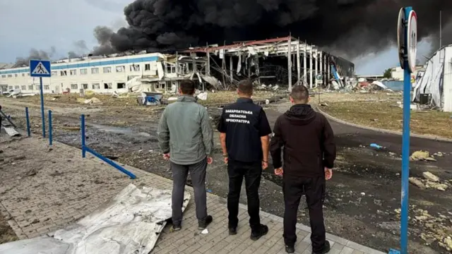 Tres hombres están de espaldas a la cámara mientras miran los escombros de un edificio que aún se quema y lanza una enorme columna de humo negro al cielo