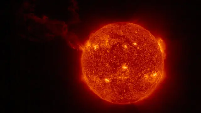 El Sol se ve como una bola roja con puntos brillantes de color amarillo en su superficie; una larga nube se extiende desde la izquierda.