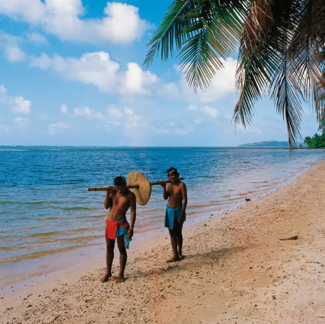 Dos hombres en la playa cargando una rai valiéndose de un palo atravezado por el hueco