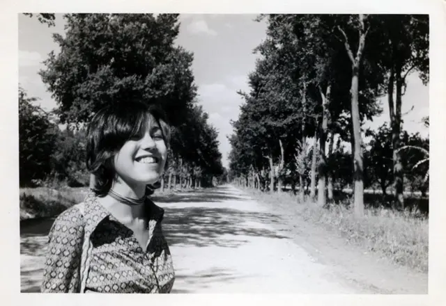 Una imagen en blanco y negro de Mariona cuando era joven. Está en una carretera bordeada de árboles y sonríe.