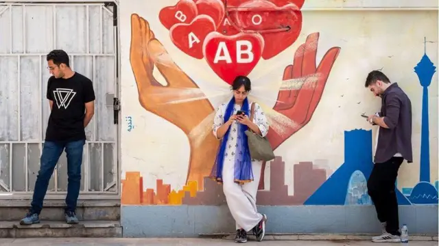 Tres personas están de pie frente a un colorido mural pintado en una pared. El mural muestra dos manos grandes, una roja y otra naranja, sosteniendo figuras con los grupos sanguíneos 