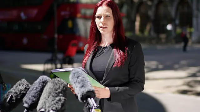 Sarah Myatt, que tiene el pelo largo teñido de rojo y viste un blazer negro sobre una blusa negra, habla con los medios mientras lee documentos en un portapapeles.