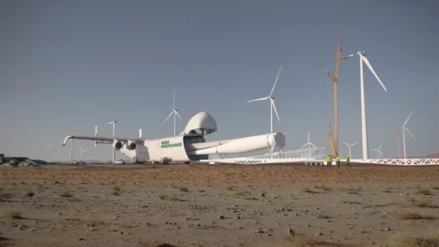 Arte conceptual de un avión blanco con el logotipo y nombres de Radia y WindRunner que está en tierra, en un parque eólico en una región desértica. La compuerta de la bodega de carga del avión está abierta y de esta salen unas enormes aspas blancas. En el fondo hay varias turbinas eólicas, unas bajo construcción