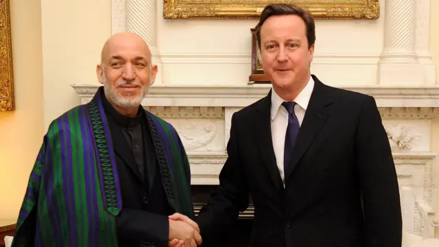 Una reunión en Downing Street en 2011 entre el primer ministro británico, David Cameron, con traje y corbata oscuros, y el presidente afgano, Hamid Karzai, quien también viste traje y camisa oscuros y un chapán bordado (abrigo o capa afgana) con rayas moradas y verdes. Se estrechan la mano frente a una chimenea de mármol y cuadros con marcos dorados.