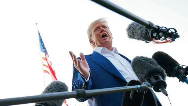 Donald Trump vestido con chaqueta de traje y camisa de cuello abierto rodeado de micrófonos. La imagen está tomada desde abajo.