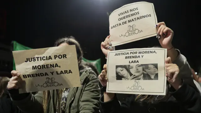 Tres manifestantes sostienen carteles durante una protesta por el asesinato de las tres mujeres argentinas. En los carteles se lee 