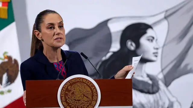 La presidenta mexicana, Claudia Sheinbaum, habla durante la sesión informativa matutina diaria. Lleva una chaqueta azul oscura y debajo una blusa que parece un huipil tradicional mexicano de color azul y rojo. En el fondo se ve una bandera mexicana y la ilustración de una mujer indígena.