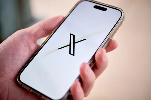 Una mano sujeta un teléfono que tiene en su pantalla la X, símbolo de la red social antes conocida como Twitter.