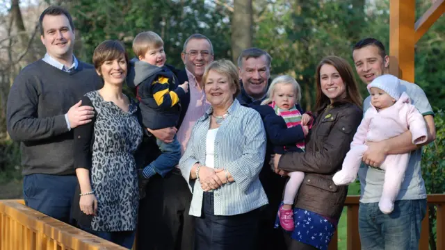 Foto de Rob, Dianne y Ronnie (en el centro) con sus dos hijos, Lloyd y Kate, sus parejas y tres nietos. Todos están de pie en un jardín, con árboles al fondo.