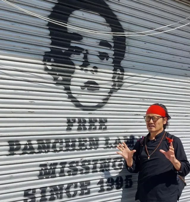 El activista y seguidor Tenzin Tsundue se encuentra frente a una imagen del Panchen Lama pintada en la persiana de una tienda.