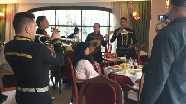 Personas almorzando en el restaurante Pozzetto: una mujer está sentada en una de las mesas mientras la rodea un grupo de mariachis.