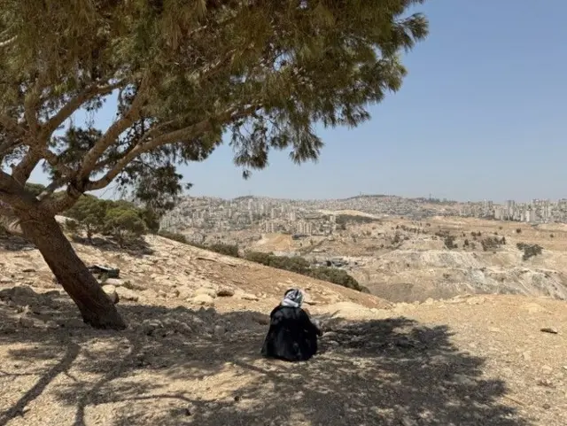 En la foto aparece Abu Emad de espaldas, a la sombra de un árbol, vestido con un traje tradicional negro y con una kufiya palestina en la cabeza, mientras mira hacia Jerusalén.