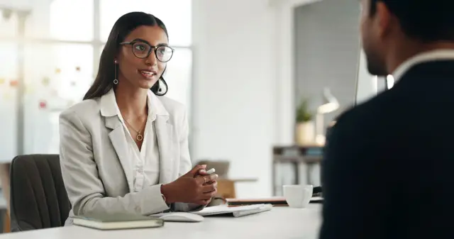 Una mujer que lleva un blazer está sentada a la mesa en lo que parece ser una oficina hablando con un hombre vestido de traje. En la mesa, se observan cuadernos, una tasa de café, un mouse y un teclado.