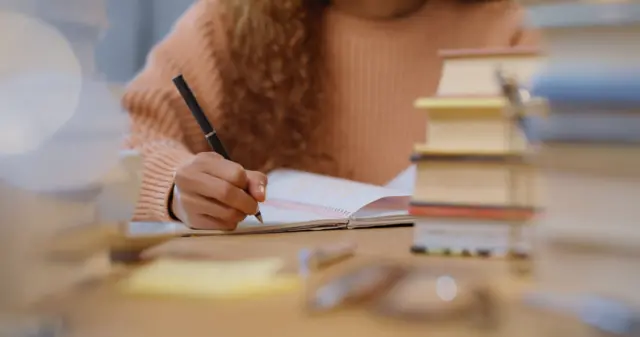 Una joven estudia, escribe en un cuaderno y la rodean varios libros. 