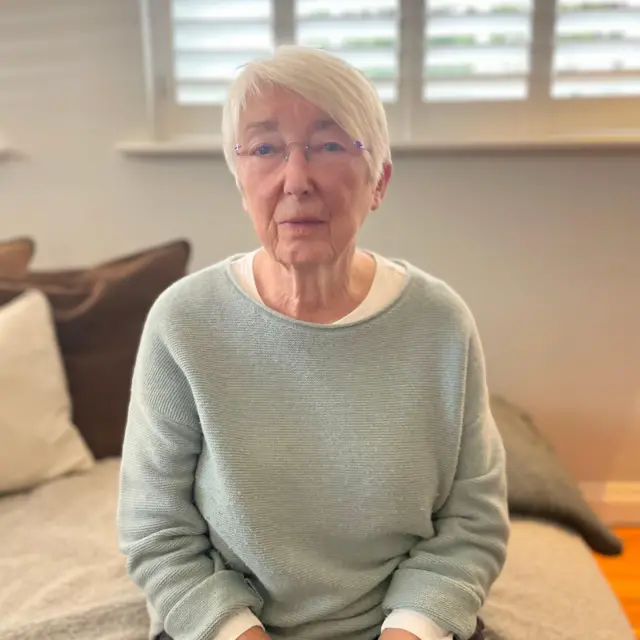 Pauline Collier sentada en una cama. Tiene el pelo corto, blanco y trae puesto un suéter azul claro   