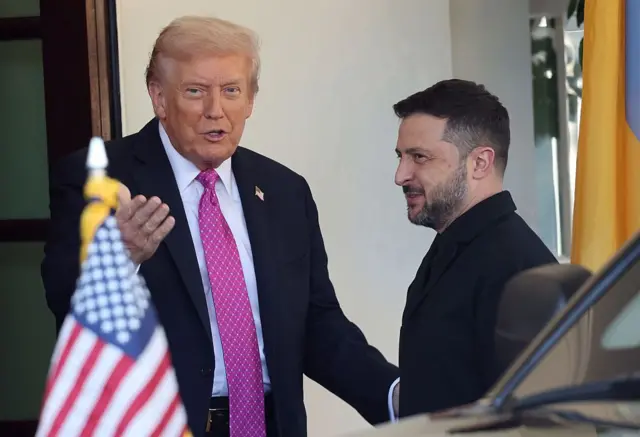 Trump saluda a Zelensky en la entrada de la Casa Blanca y dice algo que parece ser en tono jocoso. Zelensky sonríe. En primer plano se ve la bandera estadounidense en la parte delantera de un coche.