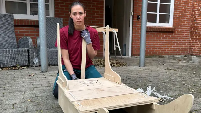 Keira, vestida con una camiseta roja y guantes protectores, está agachada detrás del trineo de madera que ha estado construyendo. Al fondo se ven un par de sillas de jardín grises y una puerta abierta que da a una casa.
