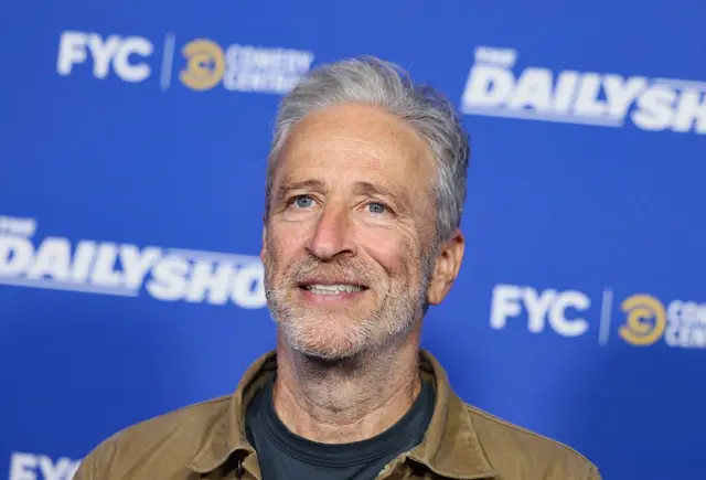 Jon Stewart, con chaqueta vaquera en tonos camel  una camiseta negra con un fondo de color azul con letras donde se promociona su programa 