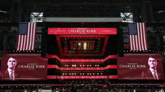 El escenario de un estadio cubierto, con grandes banderas de Estados Unidos a lado y lado y dos proyecciones de la  cara y nombre de Charlie Kirk que leen: 