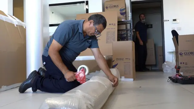 Un hombre con polo gris y pantalones oscuros arrodillado mientras envuelve con cinta adhesiva un objeto grande en plástico de burbujas. Detrás de él, hay cajas de cartón y otro hombre.  