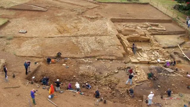 Una vista aérea de la excavación de una villa romana, que emerge de la tierra marrón. A la derecha se ven los cimientos de piedra de un edificio con dos arqueólogos en su interior. Debajo hay al menos 22 arqueólogos, de pie y de rodillas, con cubos y otros recipientes.