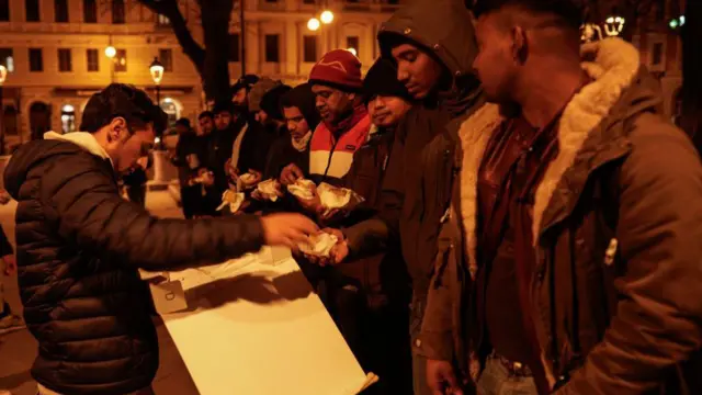 Un hombre reparte comida para llevar a una fila de hombres con abrigos y sombreros, en una plaza al aire libre en Trieste.