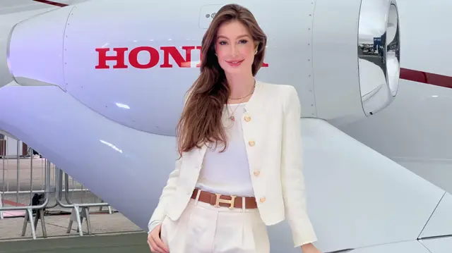 Una mujer elegantemente vestida, con pelo castaño largo y mirada confiada, frente a un jet privado