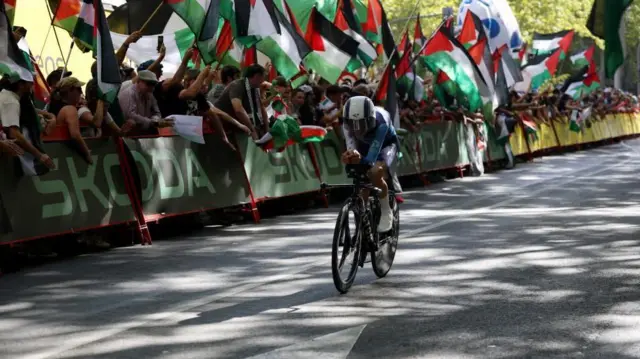 Espectadores que sostienen banderas palestinas se alinean a lo largo de la ruta de una carrera ciclista mientras un ciclista del equipo Israel Premier Tech pasa.